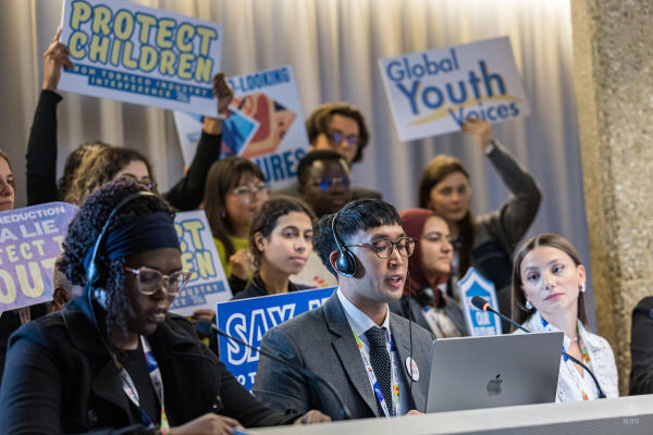 Junger Mann mit Headset, Mikrofon und Laptop verliest eine Erklärung. Dahinter halten junge Menschen Schilder mit Texten wie "Global Youth Voices" oder "Protect Children from tobacco industry interference" hoch.